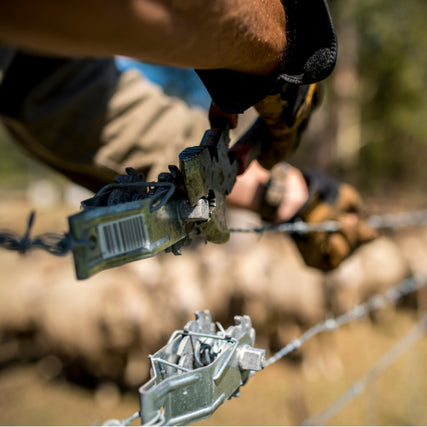 FencingClose-up of a person using pliers to tighten barbed wire on a fence. Gloves protect their hands, with a blurry rural background suggesting a farm setting.