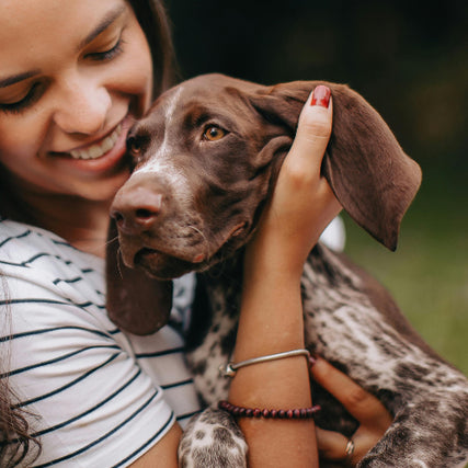 PetWoman smiling while holding a brown puppy with floppy ears. She wears striped shirt and bracelets. The scene conveys warmth and affection outdoors.
